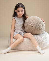 Wigiwama teddy sierkussen in beige kleur als decoratief accent. Op de bijpassende poef zit een meisje die de bal vasthoudt. Veilig, zacht en stijlvol in ieder interieur.
