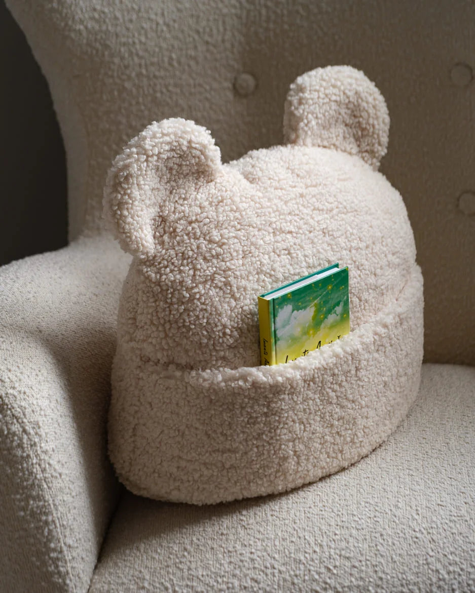 Het Wigiwama beer sierkussen in cream wit staat op een stoel met een boekje in het vakje – een functioneel én decoratief element voor rustmomenten in de kinderkamer.