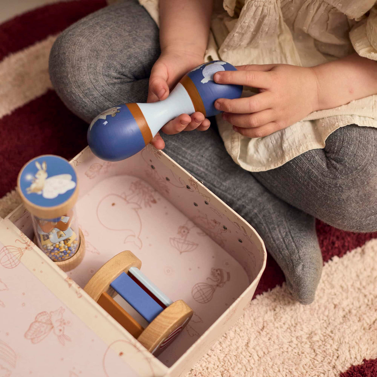 Meisje speelt met de houten maraca uit de sensorische speelgoedset van Filibabba. De rainmaker en rammelaar liggen in het koffertje, dat gemaakt is van stevig karton.