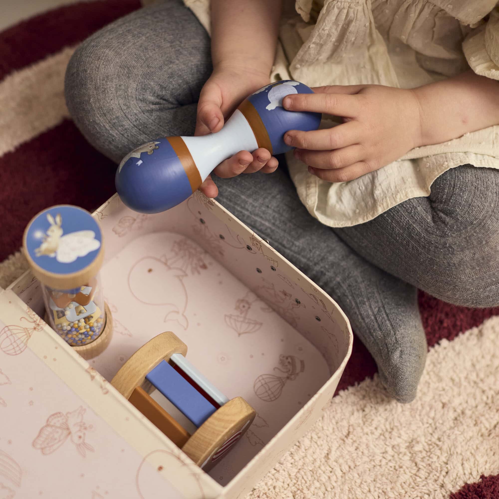 Meisje speelt met de houten maraca uit de sensorische speelgoedset van Filibabba. De rainmaker en rammelaar liggen in het koffertje, dat gemaakt is van stevig karton.