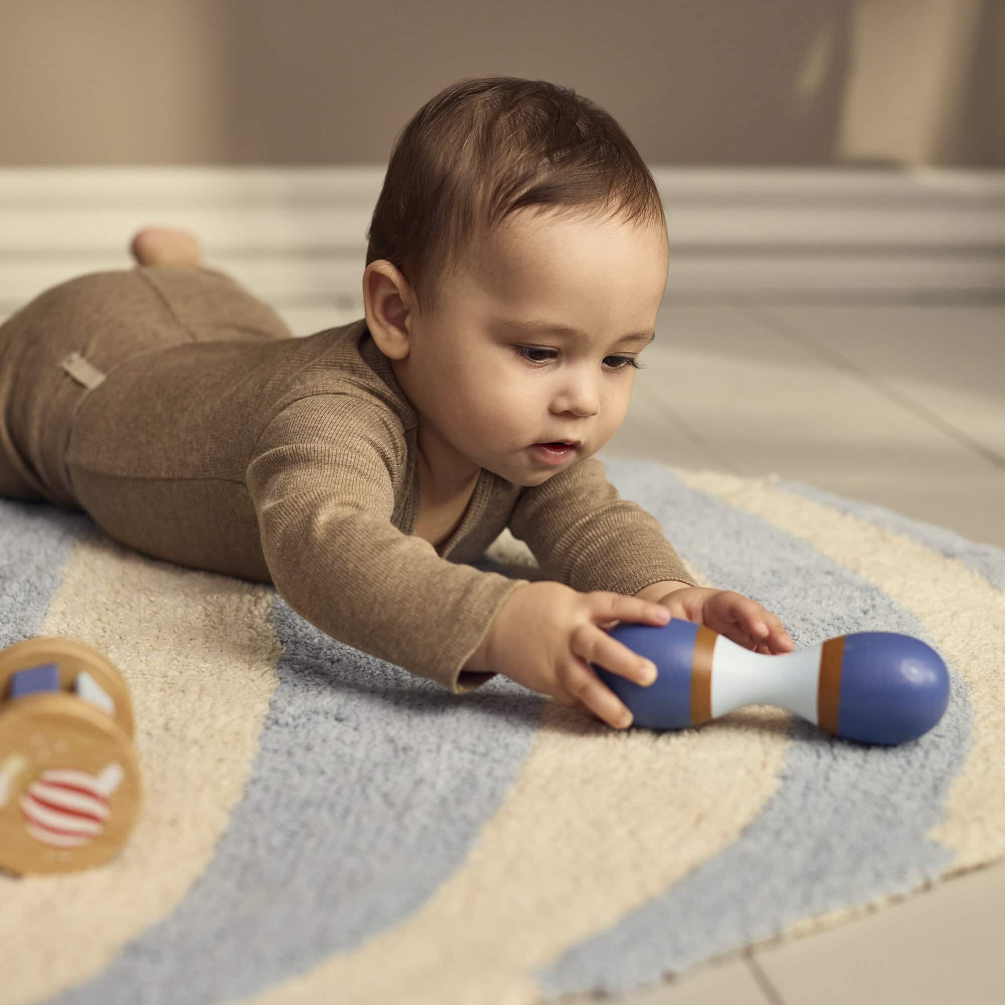 Baby speelt met de houten maraca uit de sensorische speelset van Filibabba. Het kindje ligt op een zacht speelkleed met de rammelaar aan de andere kant.