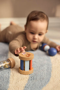 Baby reikt naar de houten rammelaar uit de sensorische speelgoedset van Filibabba. De maraca ligt naast het kindje, en de rainmaker ligt naast de gekleurde rammelaar.