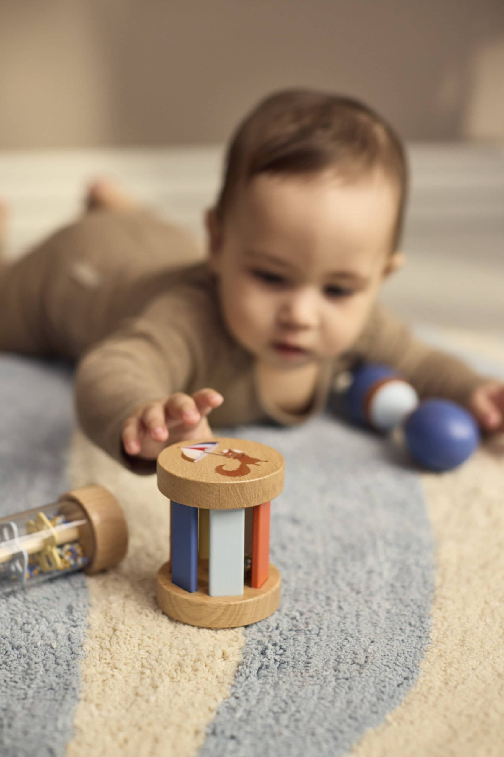 Baby reikt naar de houten rammelaar uit de sensorische speelgoedset van Filibabba. De maraca ligt naast het kindje, en de rainmaker ligt naast de gekleurde rammelaar.