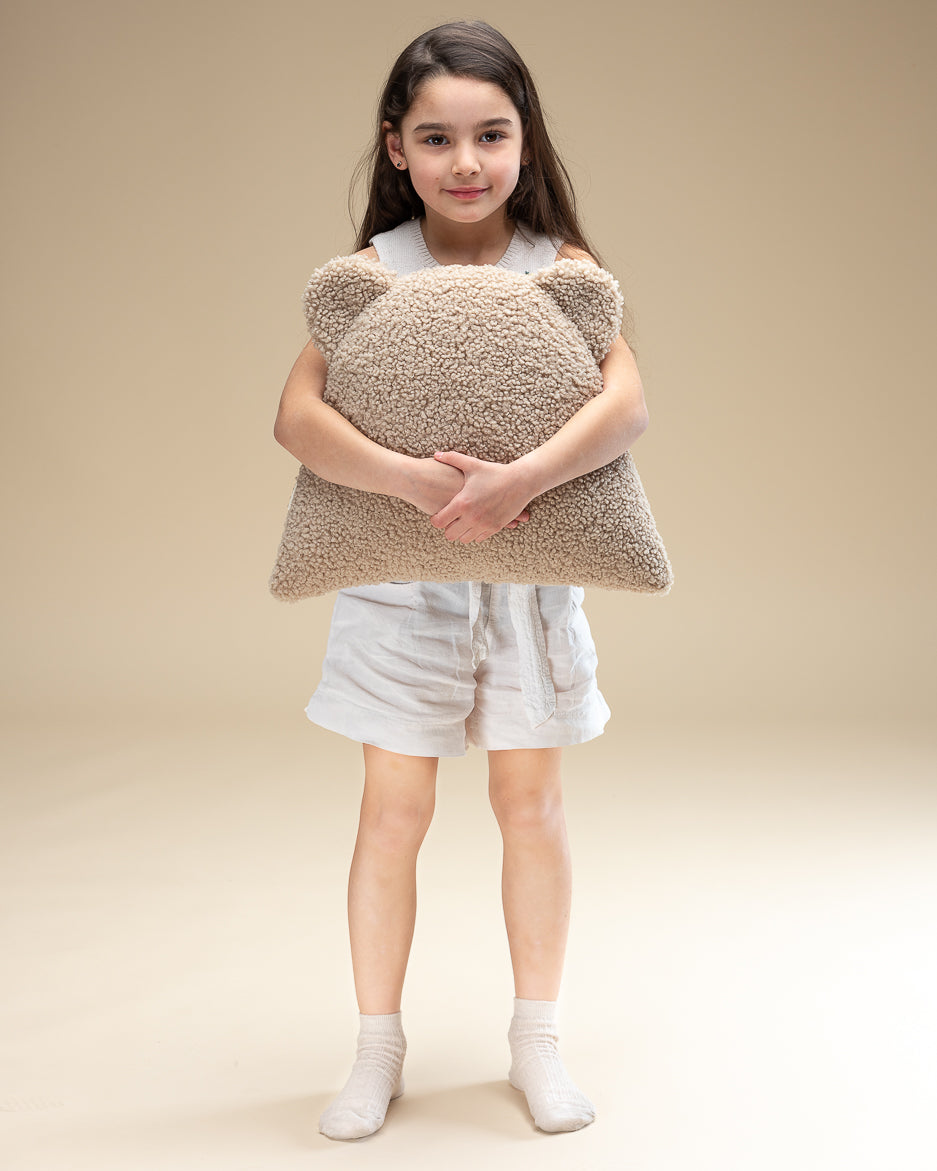Een jong meisje houdt liefdevol een teddy sierkussen in de vorm van een beertje vast. Het zachte OEKO-TEX® teddystof in biscuitkleur maakt dit kussen een veilig en knuffelbaar accessoire voor de kinderkamer.
