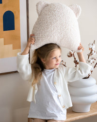 Een meisje speelt vrolijk met het schattige Wigiwama teddy beerkussen in cream. Het zachte, veilige sierkussen is een speels en functioneel accessoire voor elke kinderkamer.