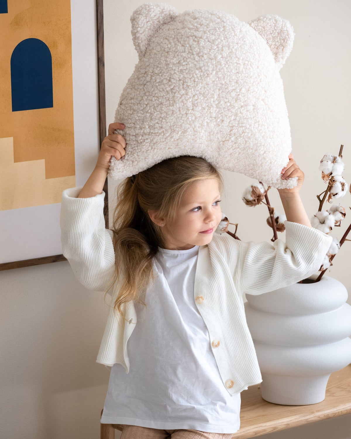 Een meisje speelt vrolijk met het schattige Wigiwama teddy beerkussen in cream. Het zachte, veilige sierkussen is een speels en functioneel accessoire voor elke kinderkamer.