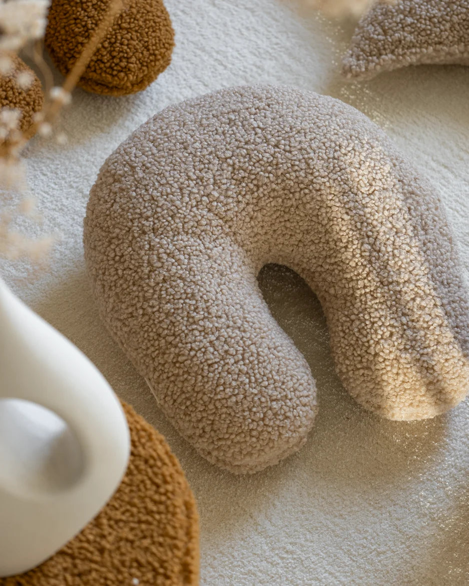 Het biscuitkleurige teddy sierkussen ligt uitnodigend op een zacht vloerkleed – een decoratieve én functionele toevoeging aan elke speelhoek of leesplek in huis.