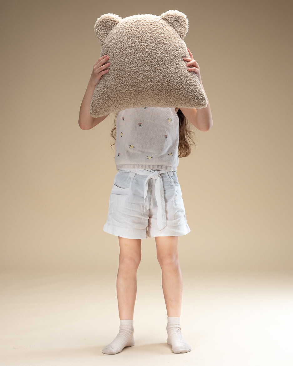 Een kind verstopt zich speels achter een beerkussen van teddystof. Met zijn schattige oortjes en zachte uitstraling is dit kussen de ideale aanvulling voor een speelhoek, bed of fauteuil in de kinderkamer.