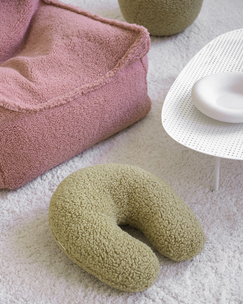 Het Wigiwama U-vorm sierkussen in natuurlijke matcha kleur ligt uitnodigend op het vloerkleed naast een roze teddy zitzak – een zachtgroene blikvanger die rust brengt in de speel- of leeshoek.