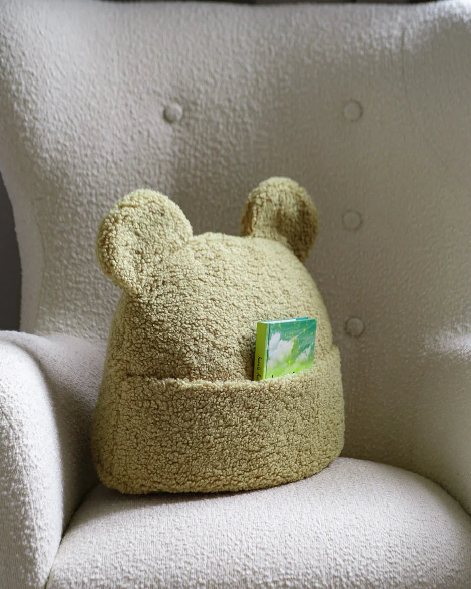 Op een comfortabele stoel ligt het Wigiwama teddy sierkussen in matcha, voorzien van een handig opbergvakje met een boekje – perfect om speelgoed of lievelingsboekjes in op te bergen.