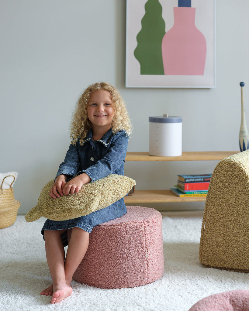 Blij meisje wat in een kinderkamer zit te poseren op de ronde roze teddy poef, op haar schoot heeft ze een okergeel teddy sierkussen. Met haar blote voetjes op een zacht wit kleed.