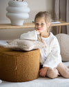 Beige stervormig teddy kussen in biscuitkleur van Wigiwama ligt op een okergele poef, naast een meisje – een stijlvol en veilig accessoire voor de kinderkamer.