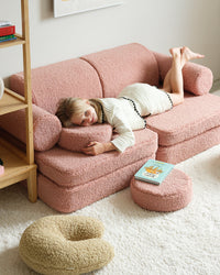 Een meisje ontspant op een teddy bank, omringd door comfortabele accessoires waaronder het Wigiwama U-vormige sierkussen in naturelle zachtgroene matcha kleur – ideaal om te relaxen, lezen of knuffelen in een gezellige kinderkamer.