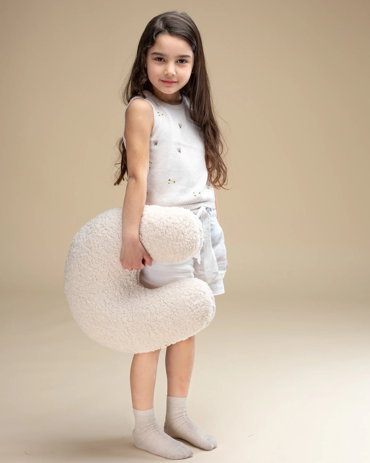 Een meisje straalt terwijl ze het zachte Wigiwama U-vorm sierkussen in cream wit vasthoudt – een tijdloze en neutrale kleur, perfect passend bij elk interieur en heerlijk om mee te knuffelen.