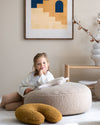 Een meisje ontspant op een grote teddy poef in een rustgevende kinderspeelhoek met het Wigiwama U-vormige kussen in okergeel maple – een sfeervolle tint die warmte en gezelligheid toevoegt aan elk interieur.