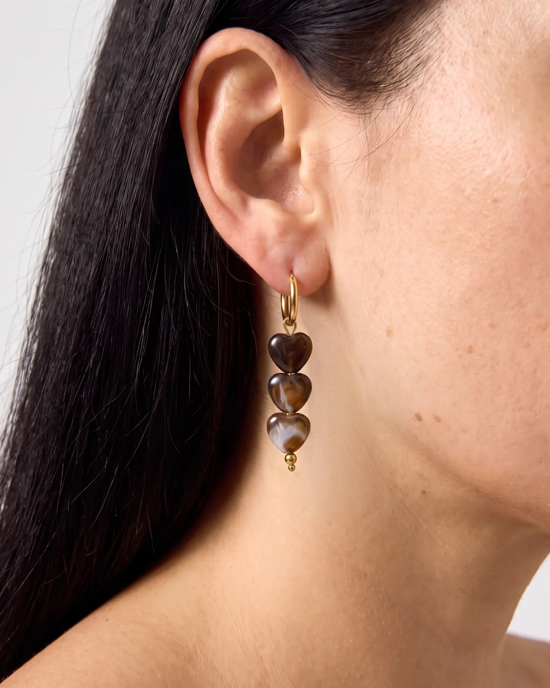 Detailfoto van oorbellen met bruine hartjes, gedragen door een vrouw met donkere haren. Hierdoor zie je de juiste grootte van de oorringetjes.