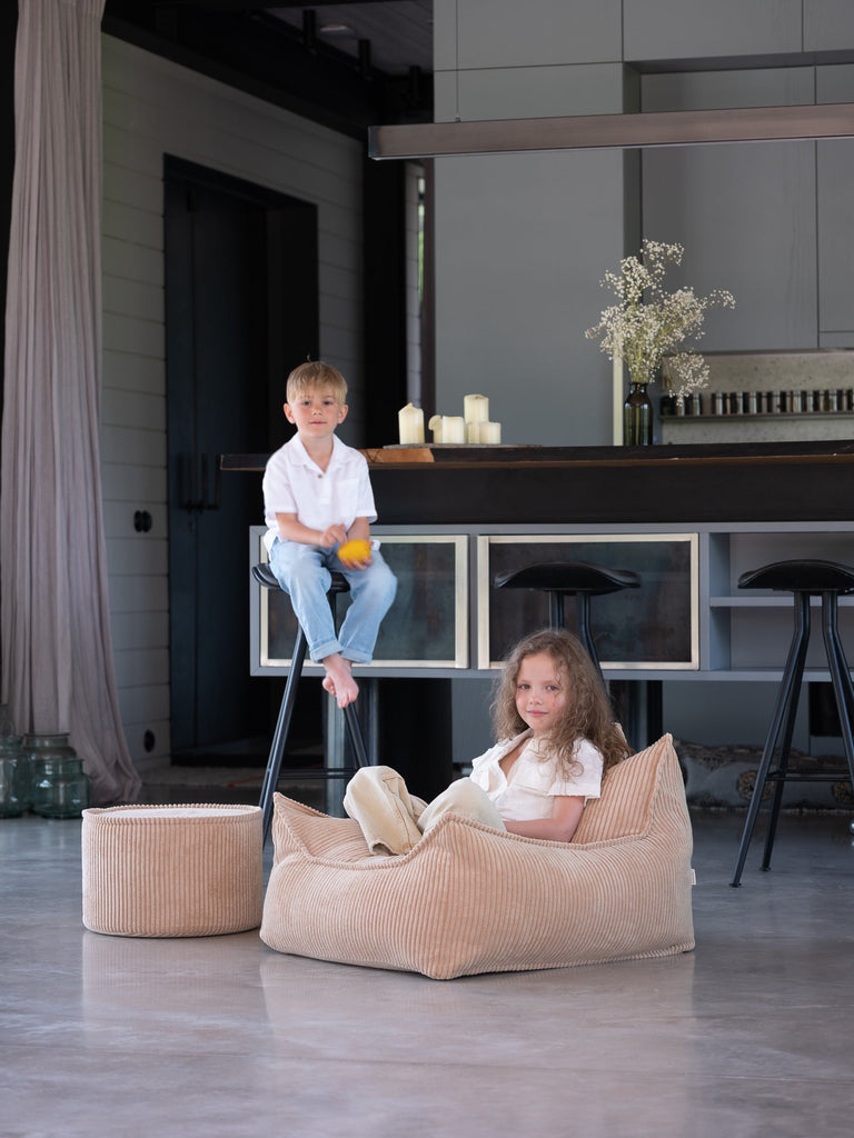 Sfeerfoto van een woonkamer waarin een jongetje op een kruk zit. Op de grond staat de beige poef en in de grote beige ribstof zitzak zit een meisje.