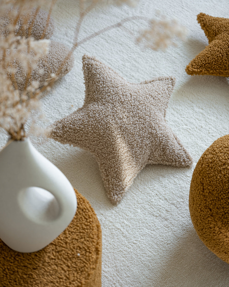 Het stervormige Wigiwama teddy kussen in biscuit – een neutrale beige tint – vormt een zacht accent op een licht kleed, ideaal voor een rustige, moderne kinderkamer.