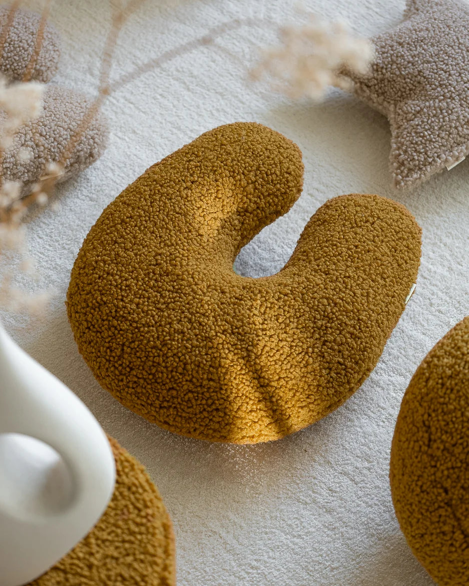 Het Wigiwama U-vormige teddy sierkussen in de kleur maple – een diepe, warme okergele tint – ligt uitnodigend op een licht vloerkleed en zorgt voor een speelse en stijlvolle touch in de kinderkamer.