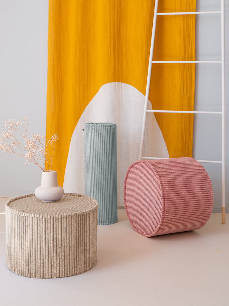 Vrolijke combinatie van een roze en beige poef en een mintgroen rolkussen. Op de beige ribstof poef staat ter decoratie een wit vaasje met takjes.