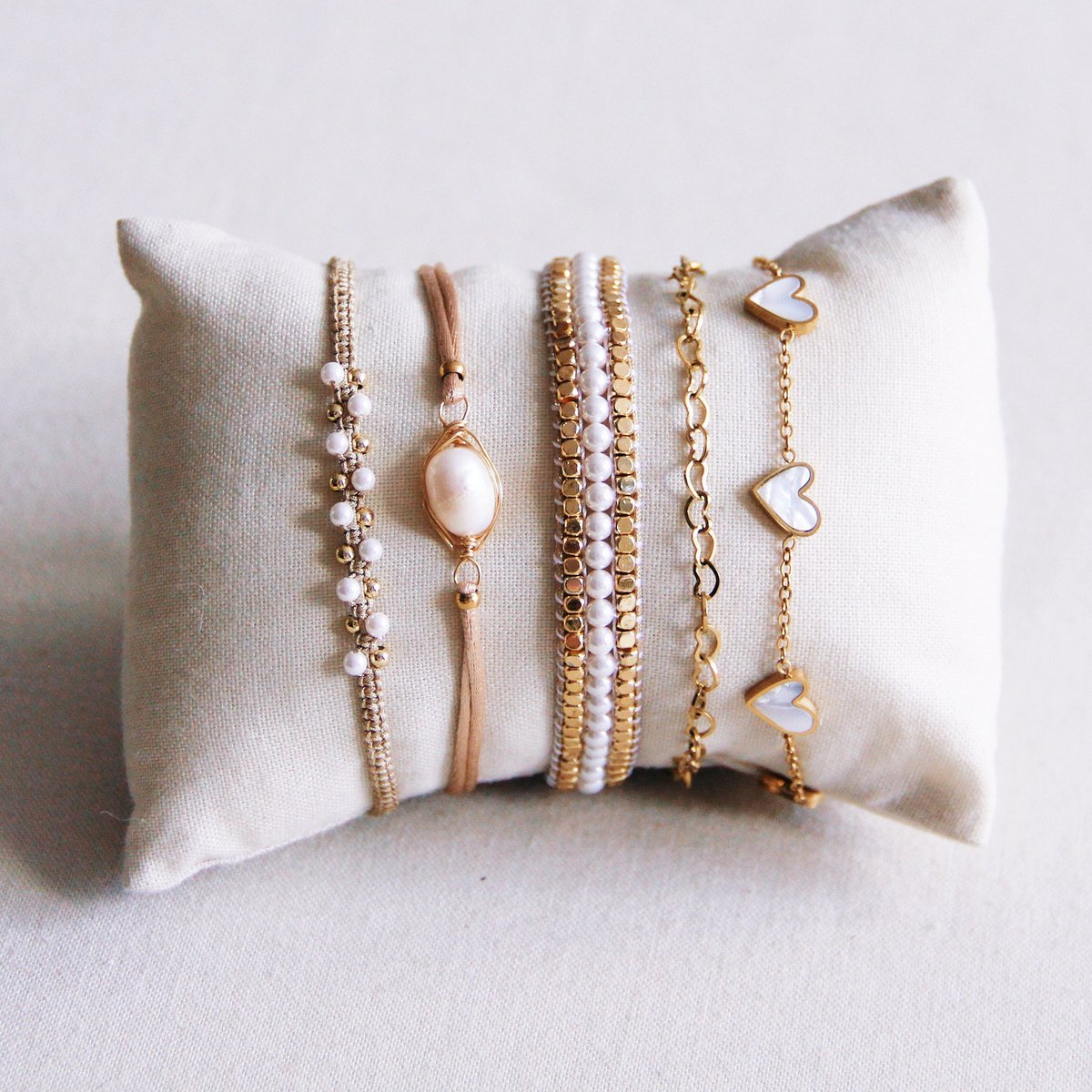 Set van vijf armbanden gepresenteerd op een linnen kussentje. Links een verstelbare beige macramé armband met witte pareltjes en gouden kraaltjes, die geschikt is voor diverse gelegenheden.