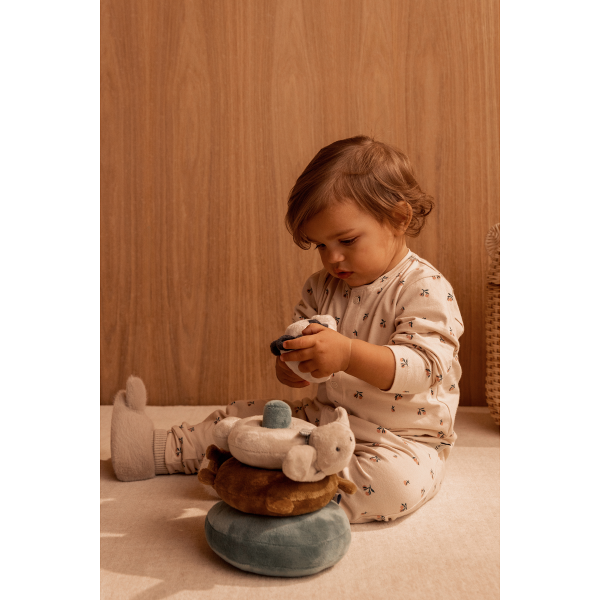 Een peuter in pyjama, spelend op de vloer, met de Beth Rabbit babyslofjes van LIEWOOD aan de voeten. Ze is aan het spelen met de Sannie Plush Stacking Tower.